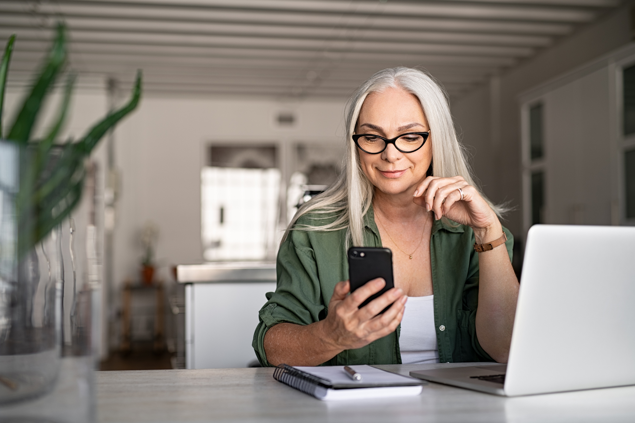 A woman sitting at a desk using her mobile phone with a notepad and laptop on top of the desk